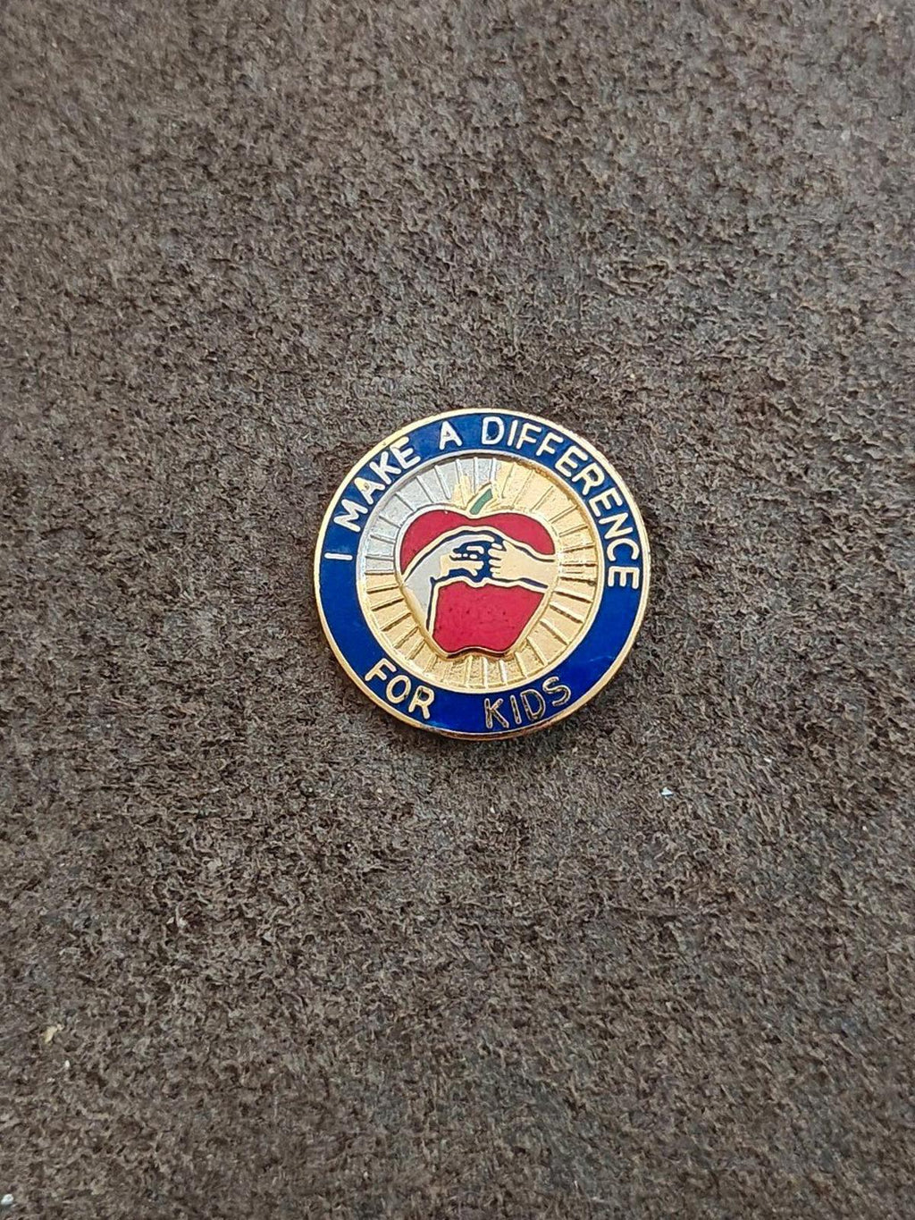 Round blue-and-gold enamel “Make a Difference for Kids” pin, apple graphic centered.