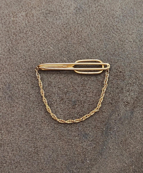 Gold-tone tie clip bar with oval hinge detail and chain on gray background