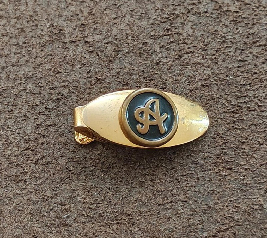 Gold-tone tie clip with blue-black circular monogram emblem centered on face