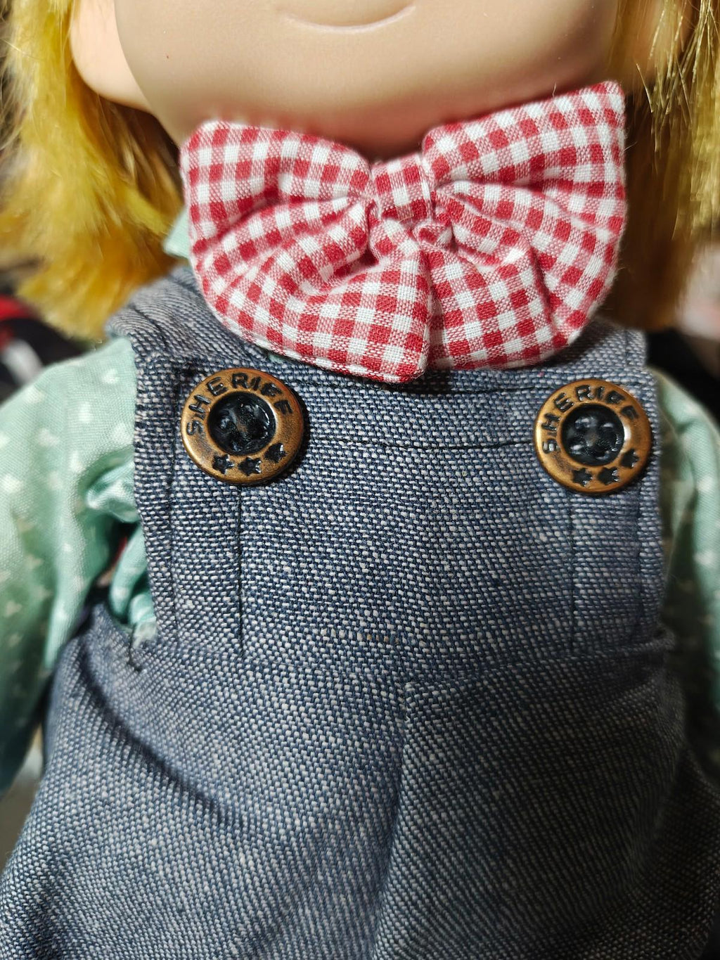 Boy doll outfit close-up: red check bow tie and blue denim overalls with brass buttons.