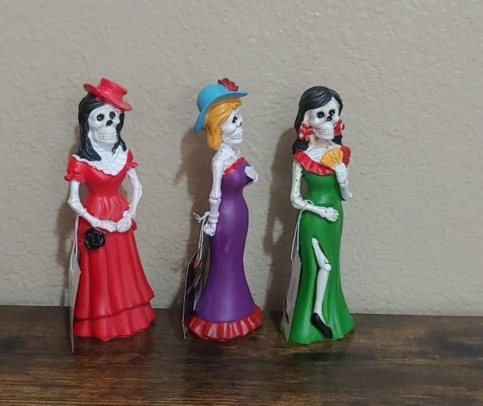 Three Catrina figurines standing together, red, green, and purple dresses