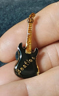 Gold-tone guitar enamel pin with black front and Beatles-style script, held on a fingertip