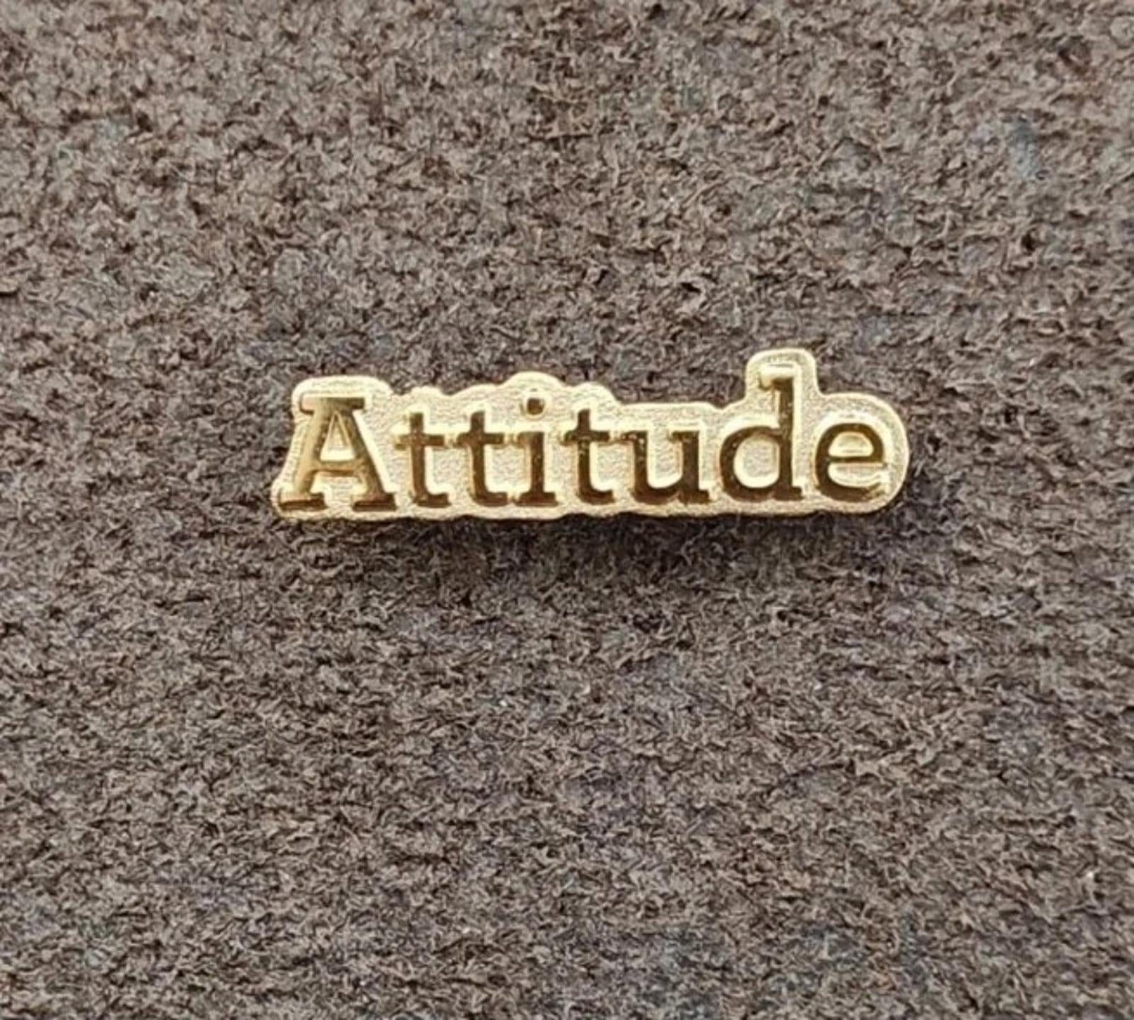 Gold tone lapel pin with raised "Attitude" lettering on gray textured background