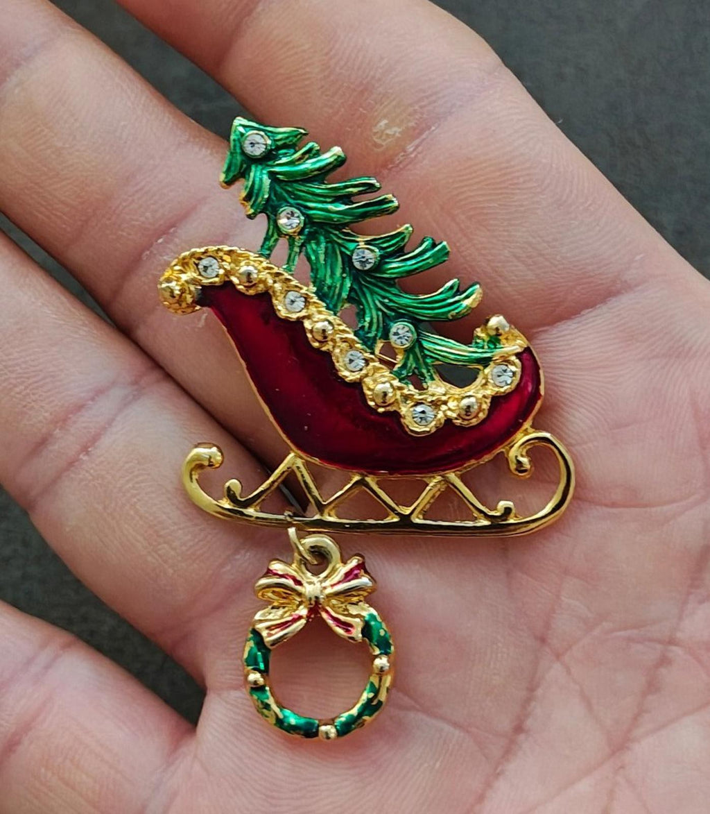 Close-up of red sleigh enamel pin with green Christmas tree, gold runners, and wreath dangle.