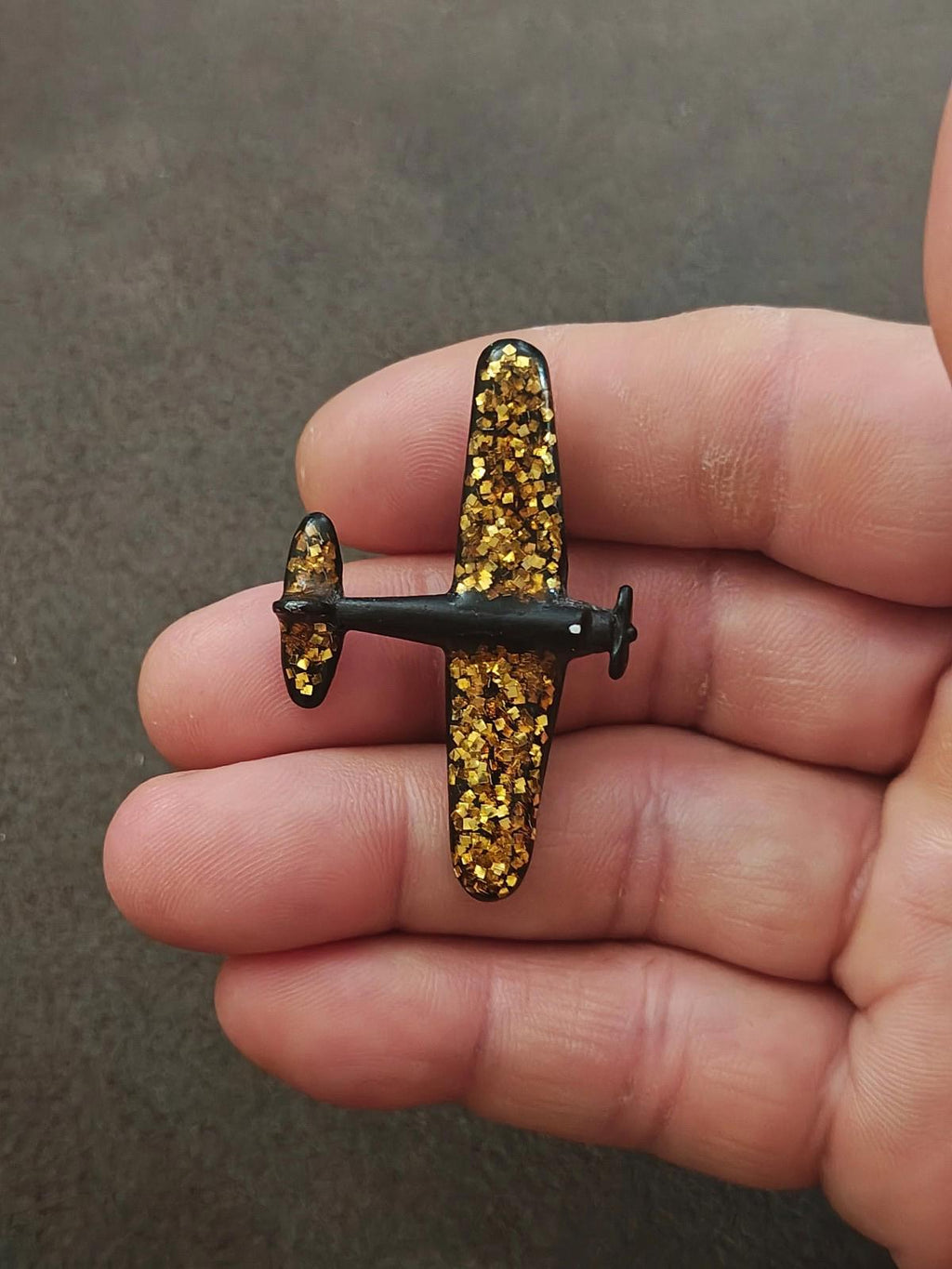 Gold glitter airplane brooch with dark posts and end caps, held in hand