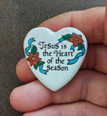 White ceramic heart pin with colorful text: “Jesus is the Heart of the Season.”