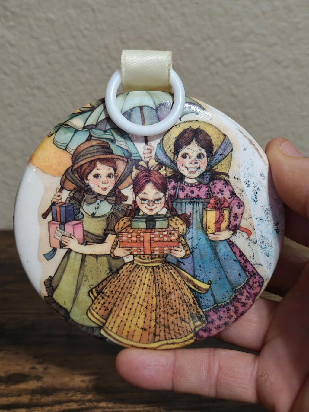 Round illustrated ornament with three girls holding a toy cart, cream loop handle