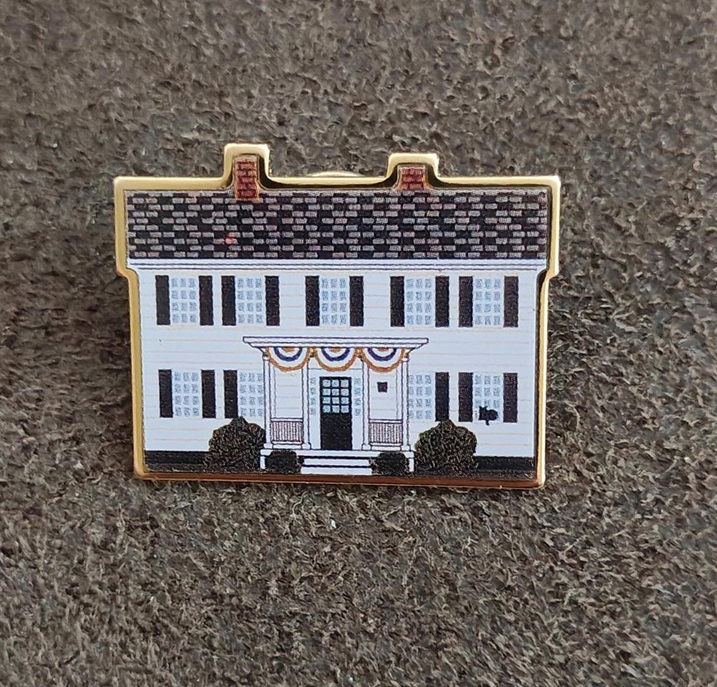 Gold-edged enamel pin on gray fabric, featuring white building facade and dark shrubs