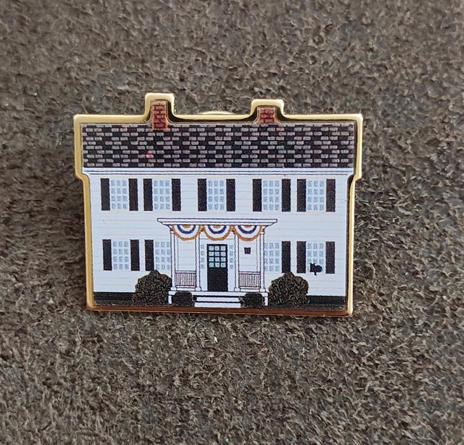 Gold-edged enamel pin on gray fabric, featuring white building facade and dark shrubs