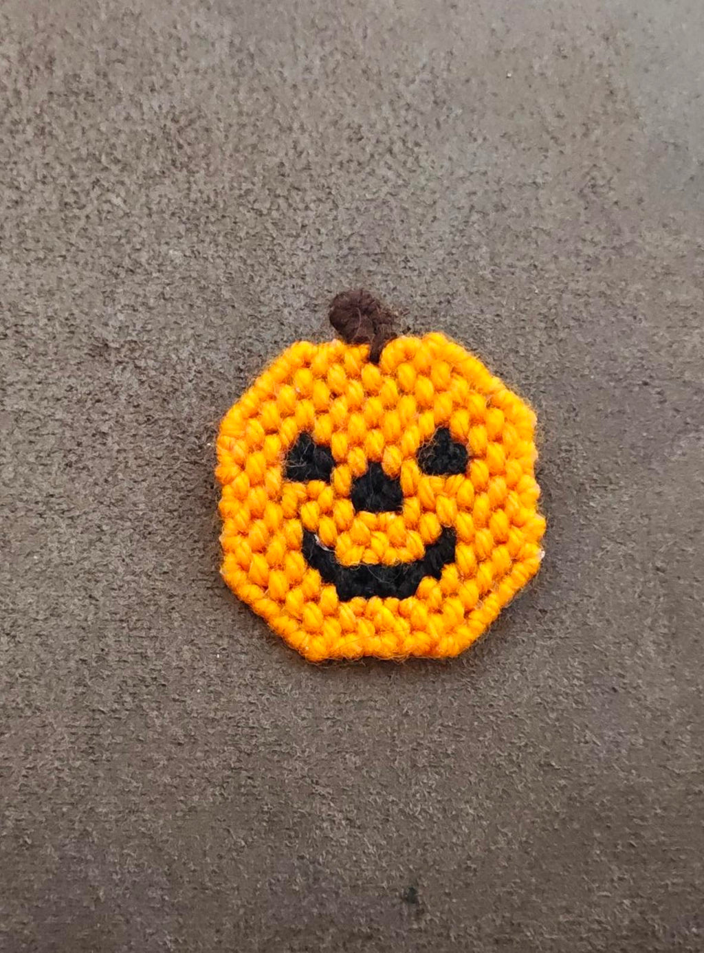 Orange knitted jack o' lantern face brooch with black eyes, nose, and grin.