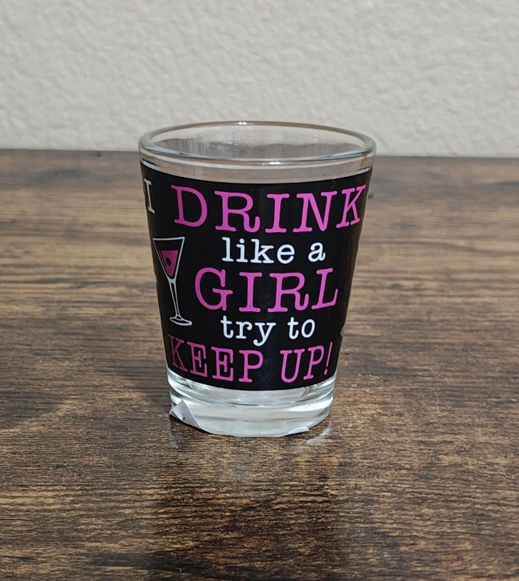Clear shot glass on table with purple “DRINK like a GIRL try to KEEP UP!” design