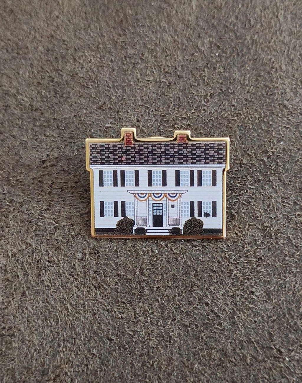 Enamel pin of a white building with blue windows and gold outline on gray fabric
