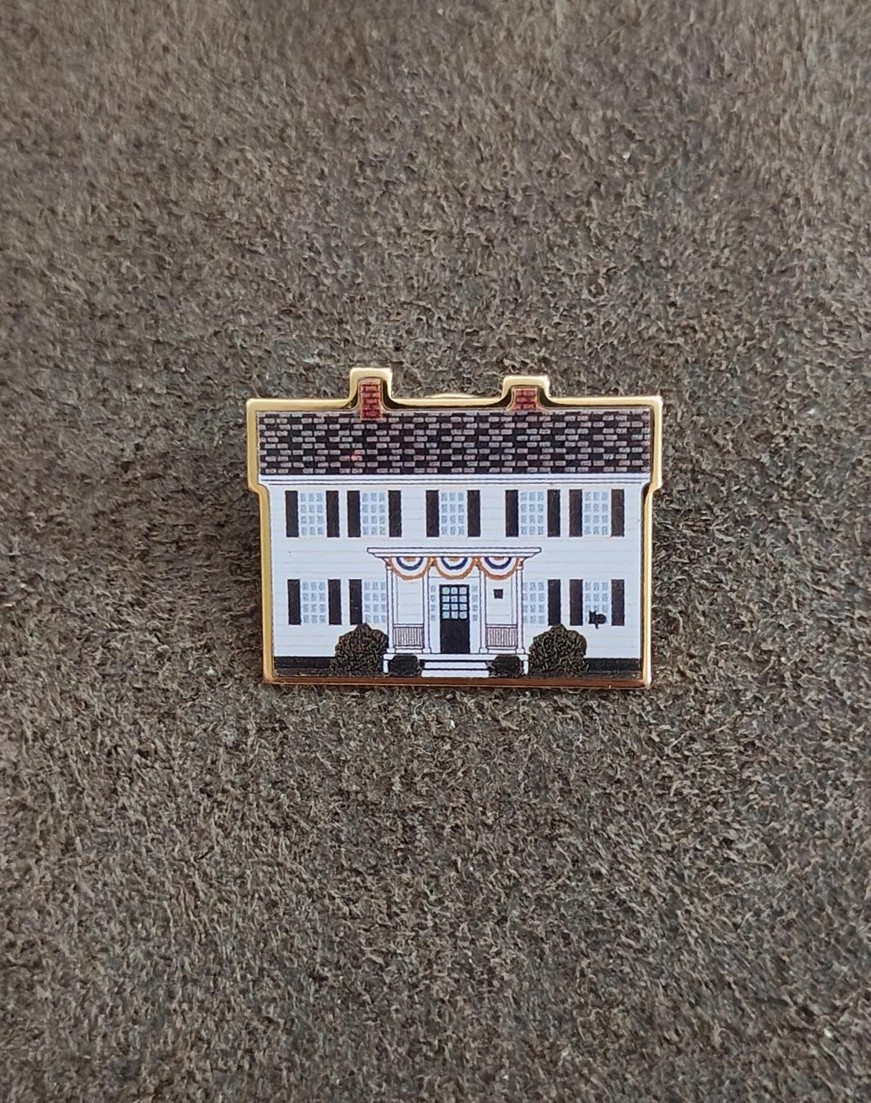 Enamel pin of a white building with blue windows and gold outline on gray fabric