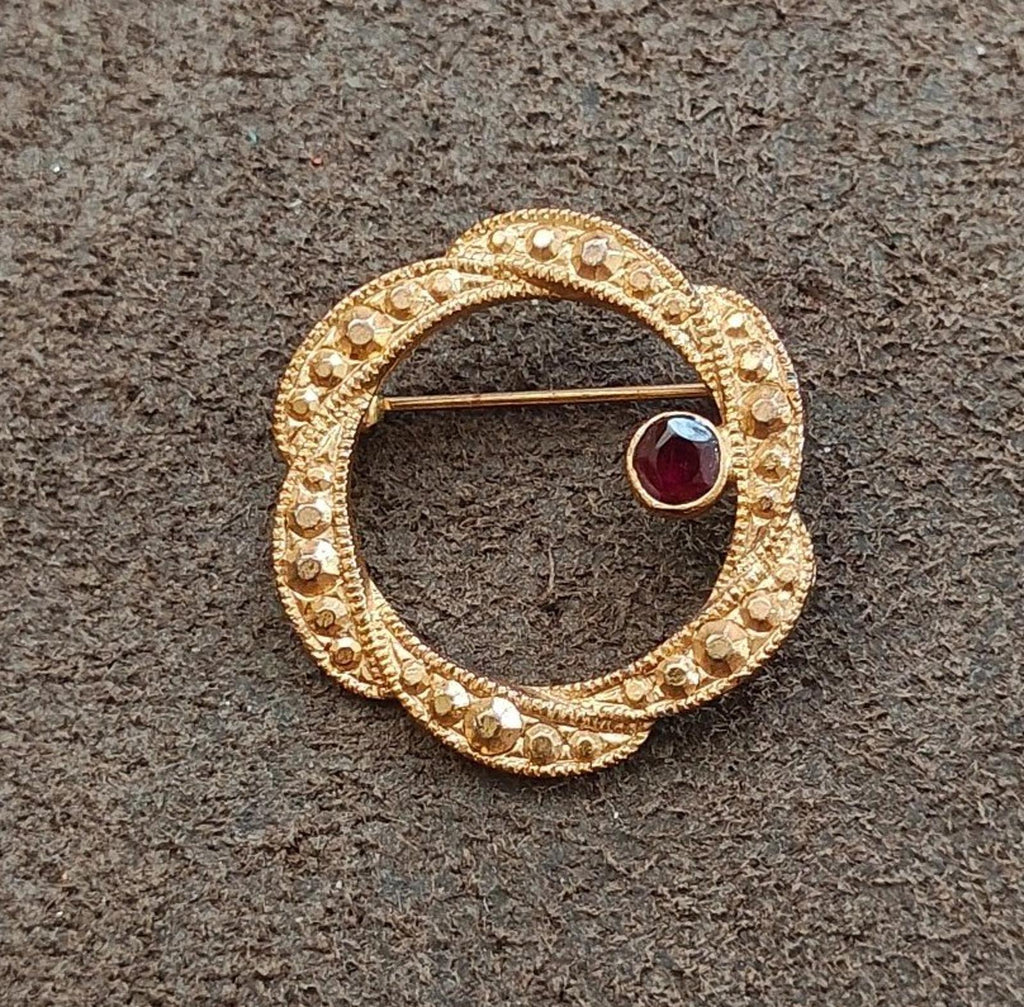 Gold tone circular brooch with dark red garnet cabochon and engraved beaded border.