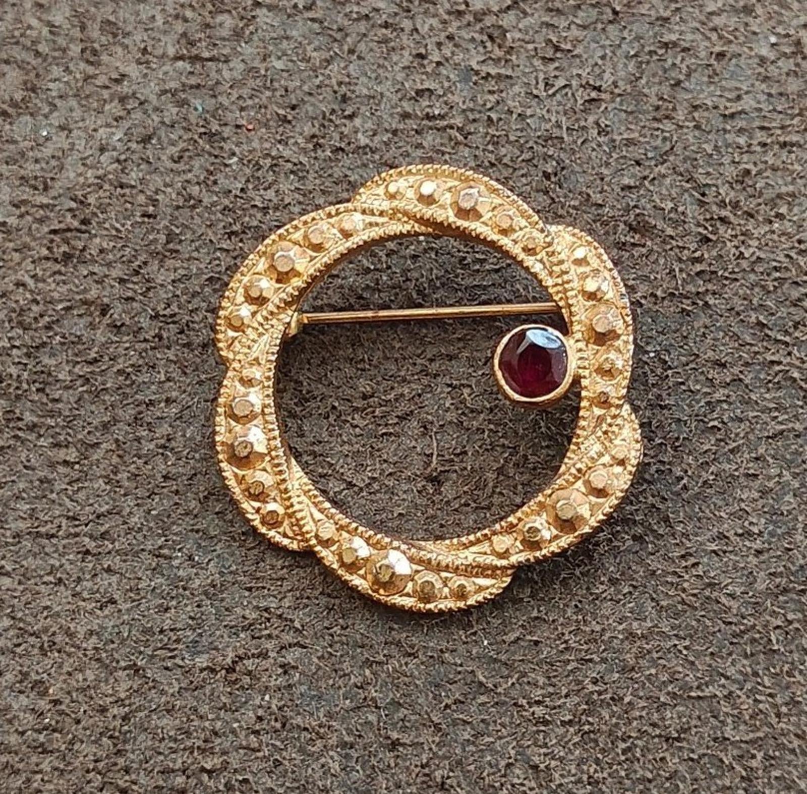 Gold tone circular brooch with dark red garnet cabochon and engraved beaded border.