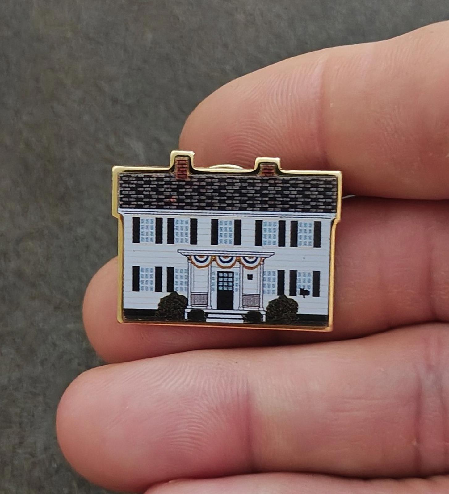 Enamel pin front held by fingers, showing white facade, blue windows, and gold border