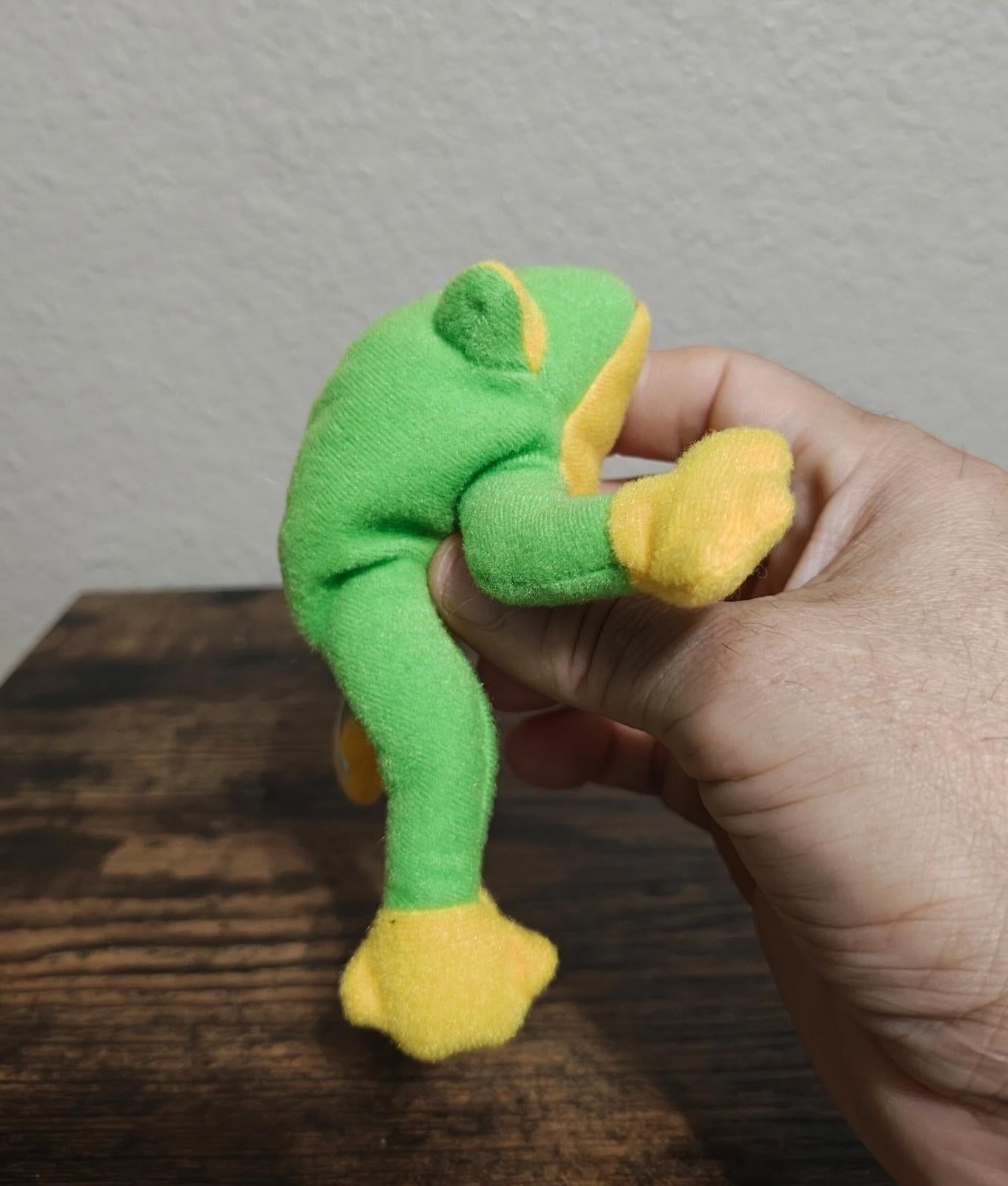 Green frog beanbag plush with yellow underside and yellow feet, held by hand