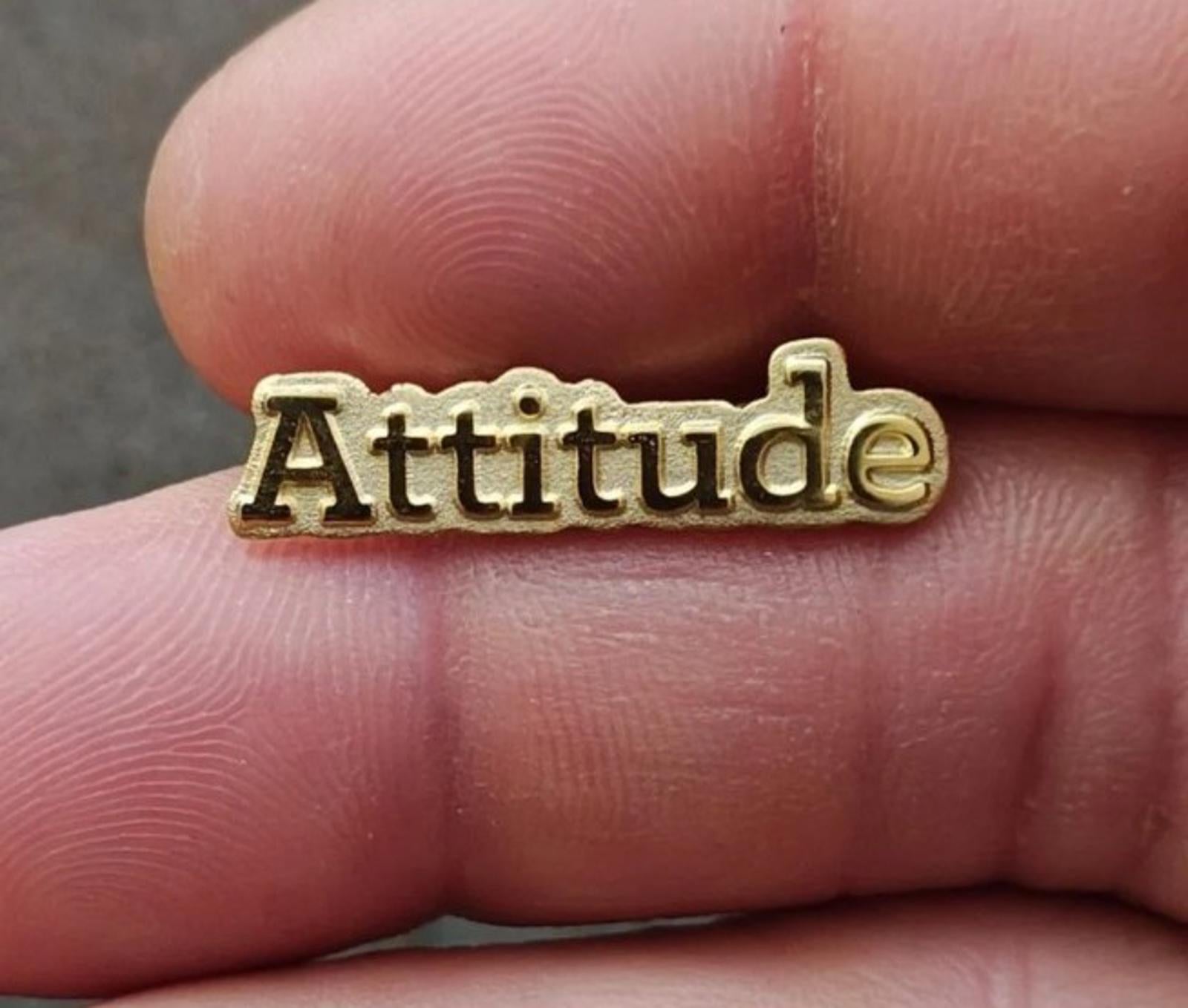 Gold tone lapel pin with raised "Attitude" lettering, close-up on fingers