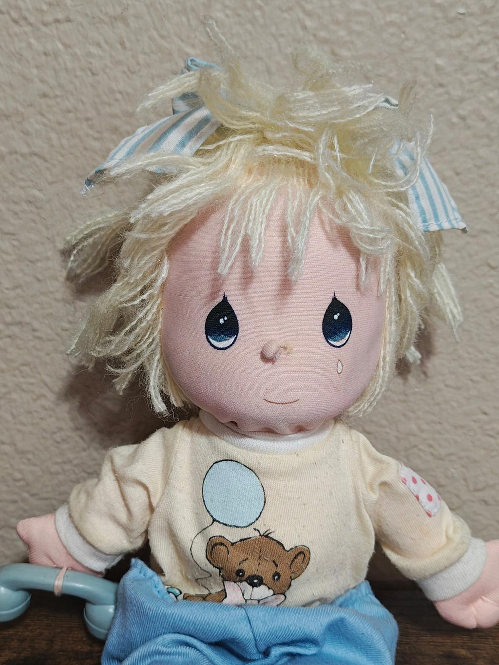 Boy doll plush close-up with blond yarn hair, cream shirt with bear graphic, and blue overalls