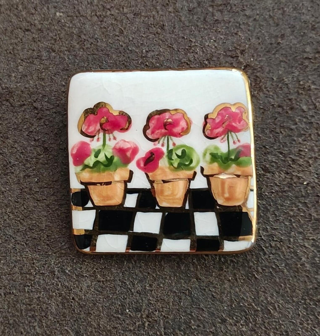 Square glazed ceramic brooch showing three pink flowers in pots on black-and-white checker base