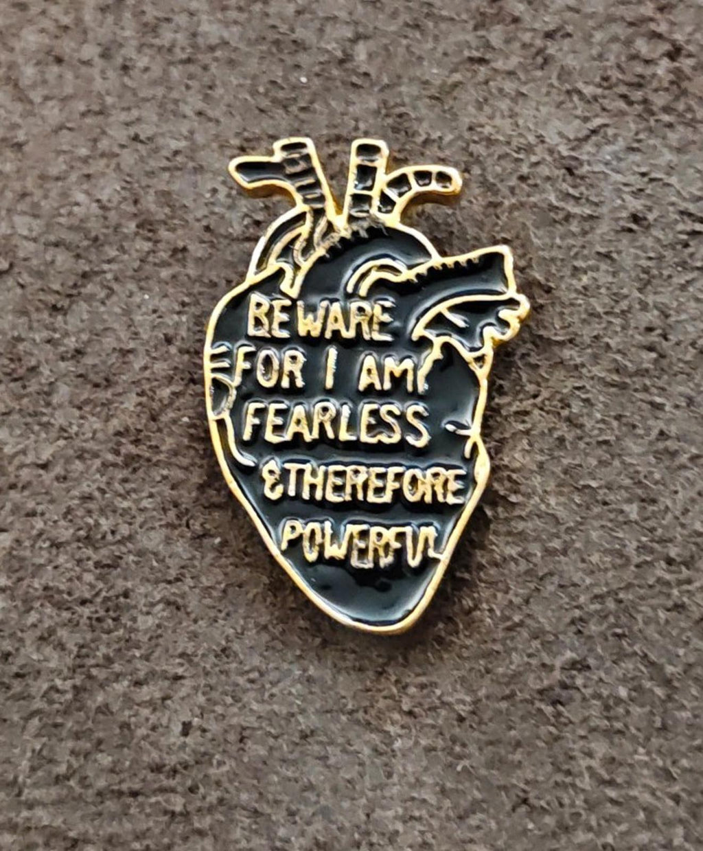 Gold-tone anatomical heart enamel pin with black enamel warning text on gray surface