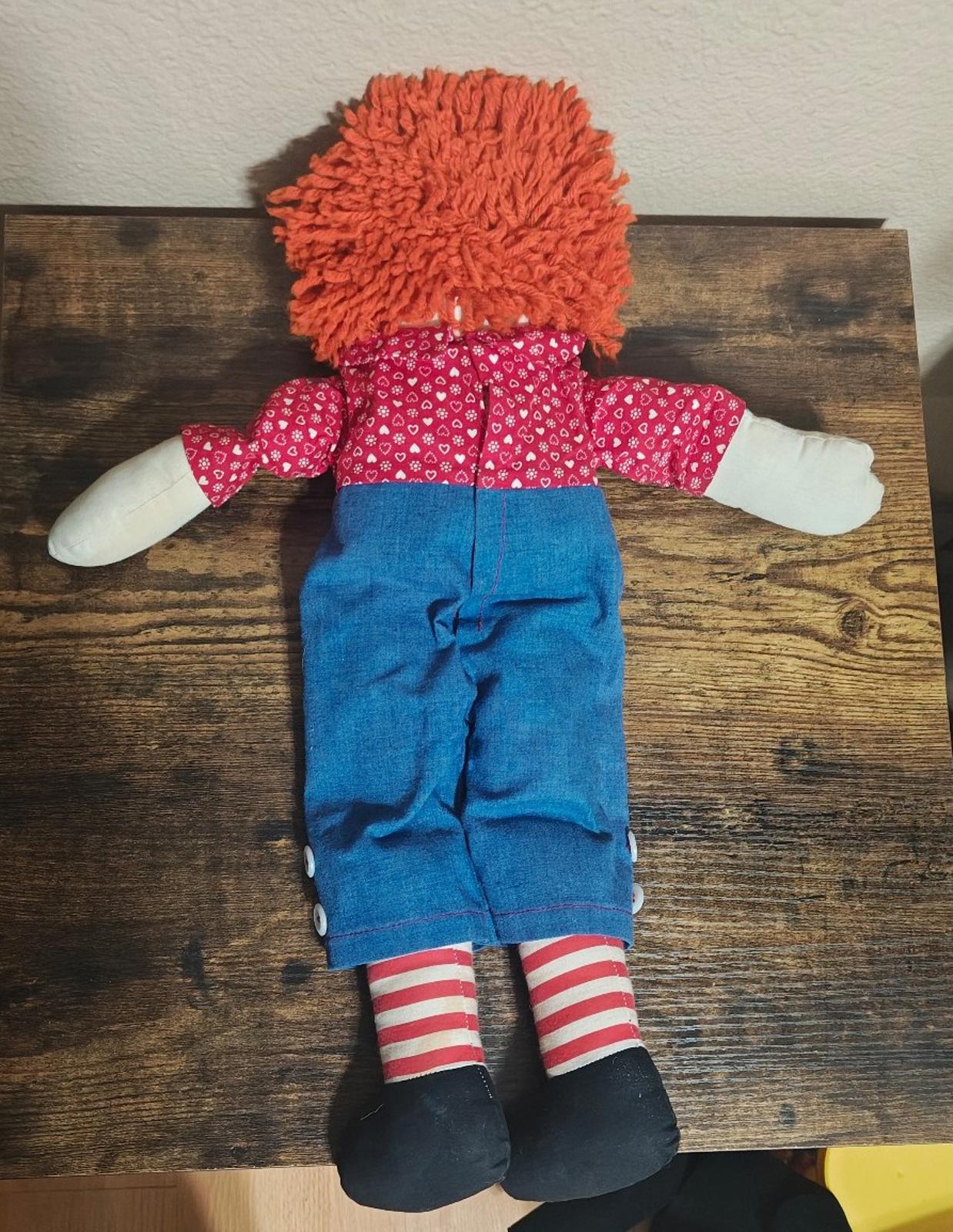 Handmade rag doll back view with orange yarn hair and red heart-pattern shirt over blue overalls.