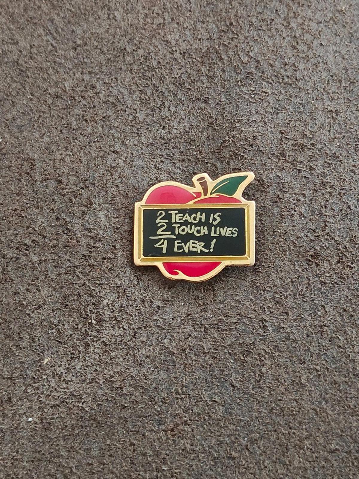 Gold-tone apple teacher pin with red apple, green leaf, and black text panel on gray surface
