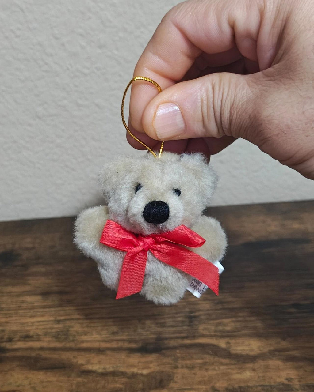 Beige mini plush teddy bear ornament with red ribbon bow and gold hanging loop, close-up