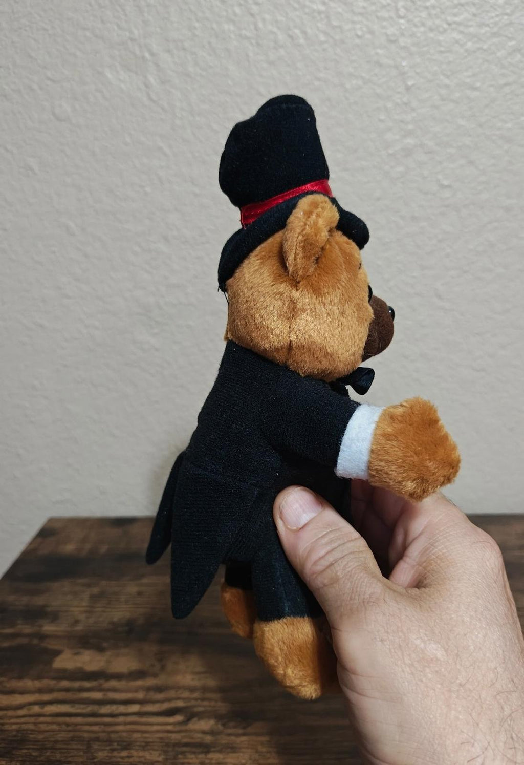 Brown plush bear turned sideways in black tuxedo with red vest and top hat.