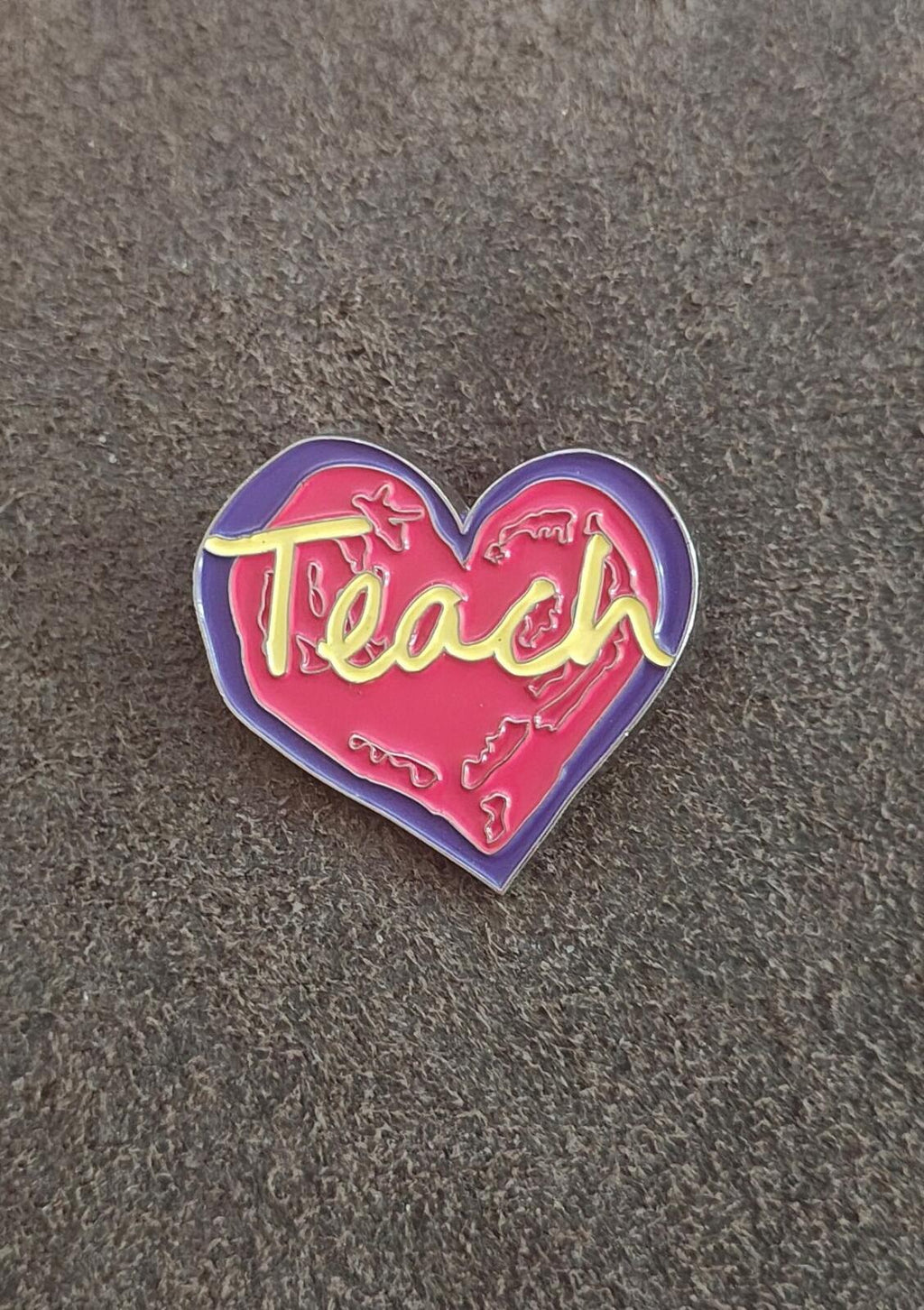 Enamel heart pin on gray fabric, purple border with red “Teach” and yellow accents.