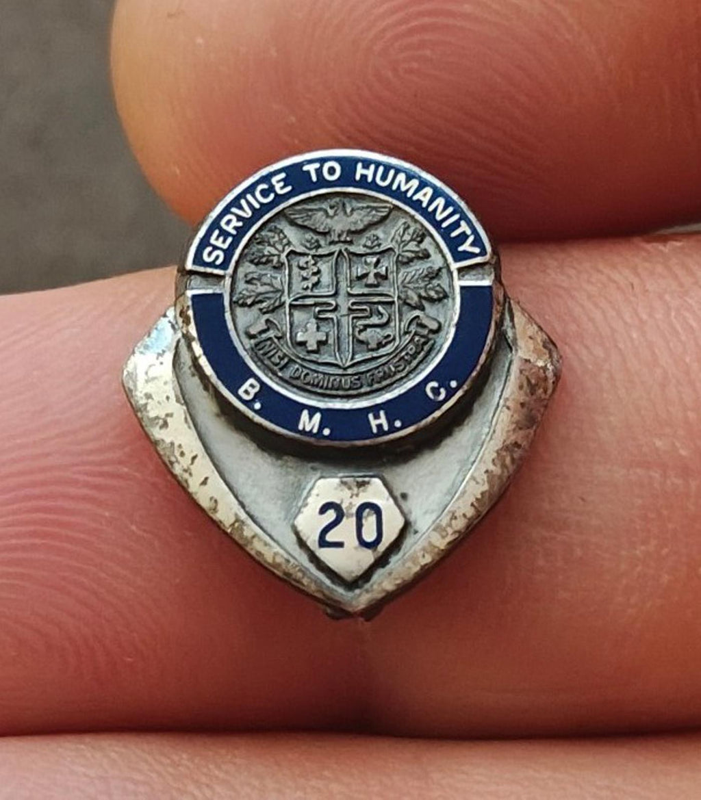Silver enamel service award pin with shield medallion, “Service to Humanity,” and “20”.