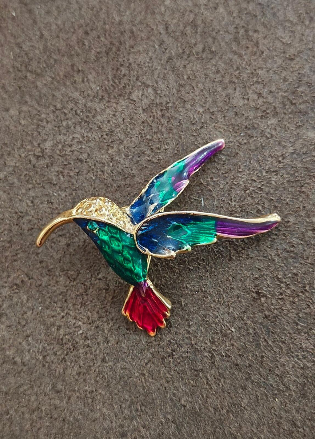 Gold tone hummingbird enamel pin on gray felt, green and purple wings with red tail