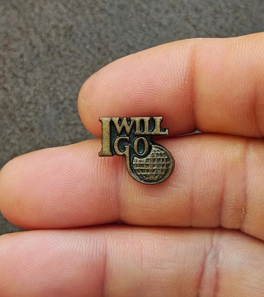 Gold tone "I WILL GO" ministry pin with globe-style emblem, close-up in hand