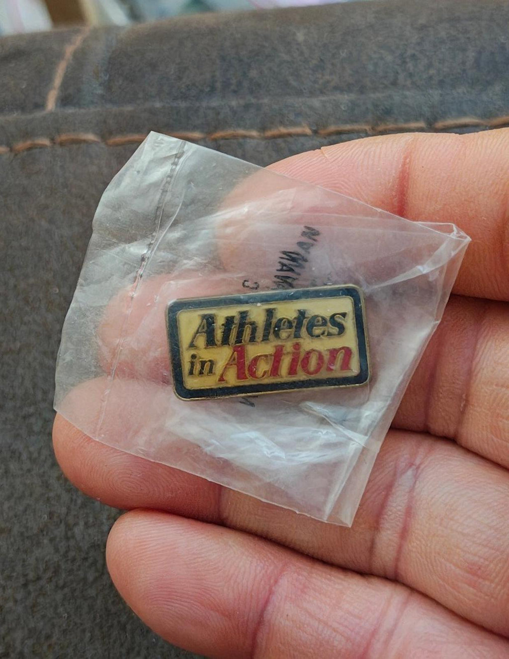 Gold-tone enamel pin reading "Athletes in Action" in clear plastic bag, held up