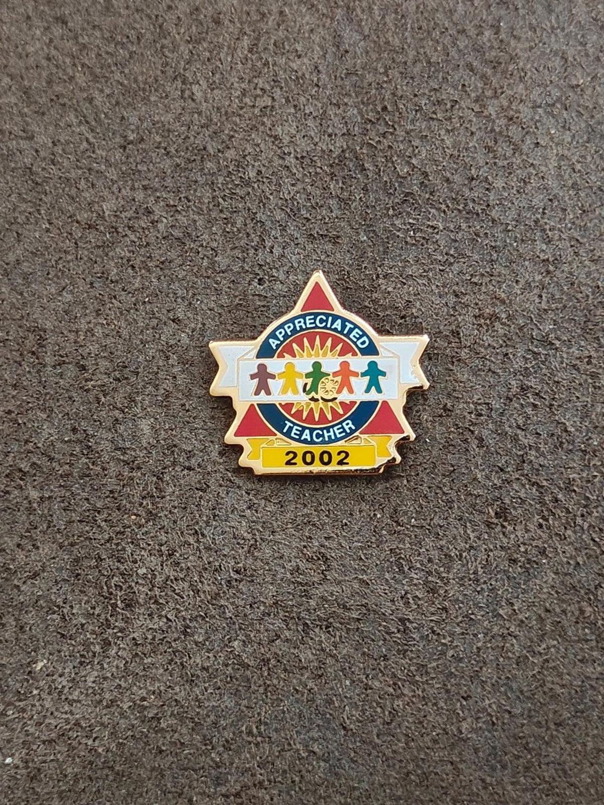 Teacher enamel pin standing on gray surface, gold-tone outline and “2002”
