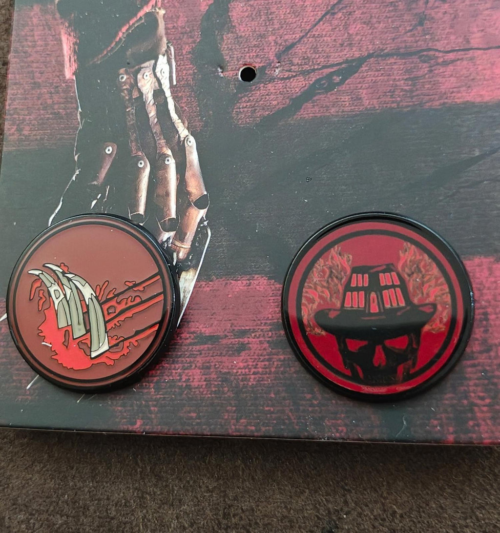 Close view of two round enamel pins: left glove emblem and right skull emblem in red/black