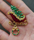 Red sleigh enamel pin with green Christmas tree, gold details, and dangling wreath charm.