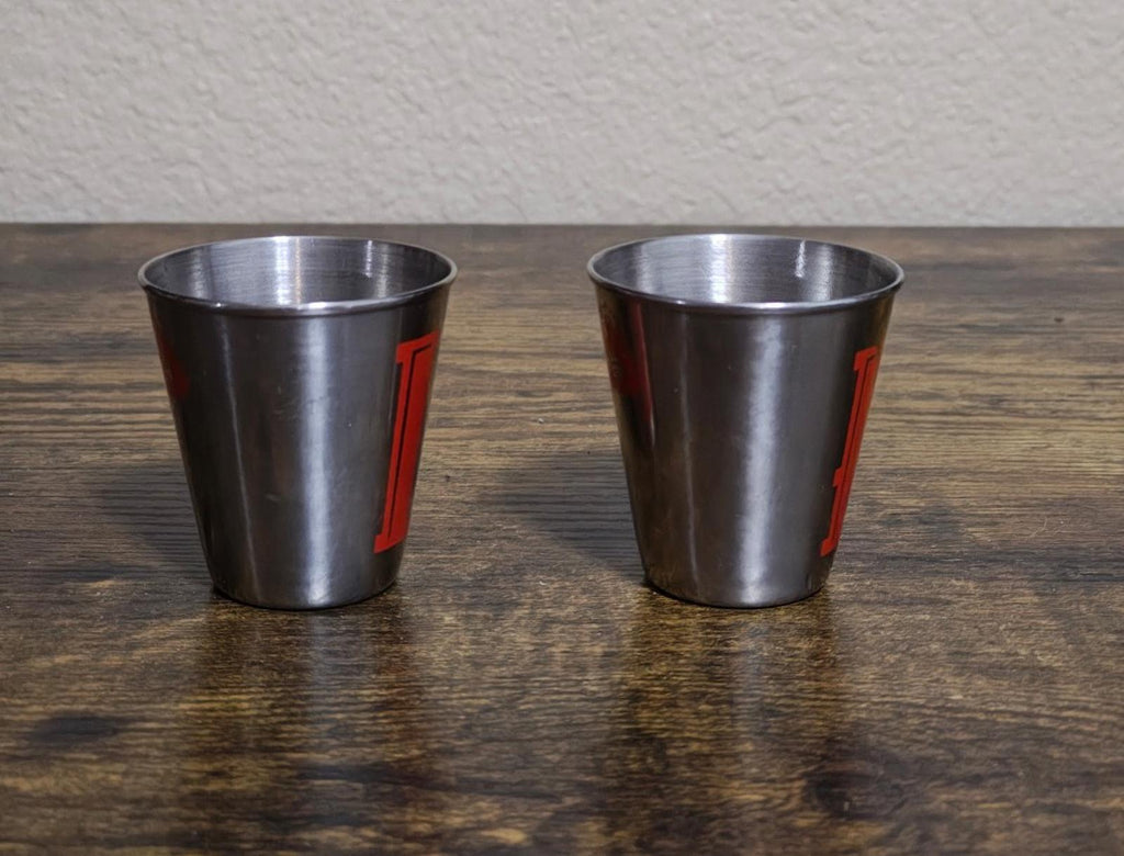 Pair of stainless steel shot glasses shown from the side with red vertical letters