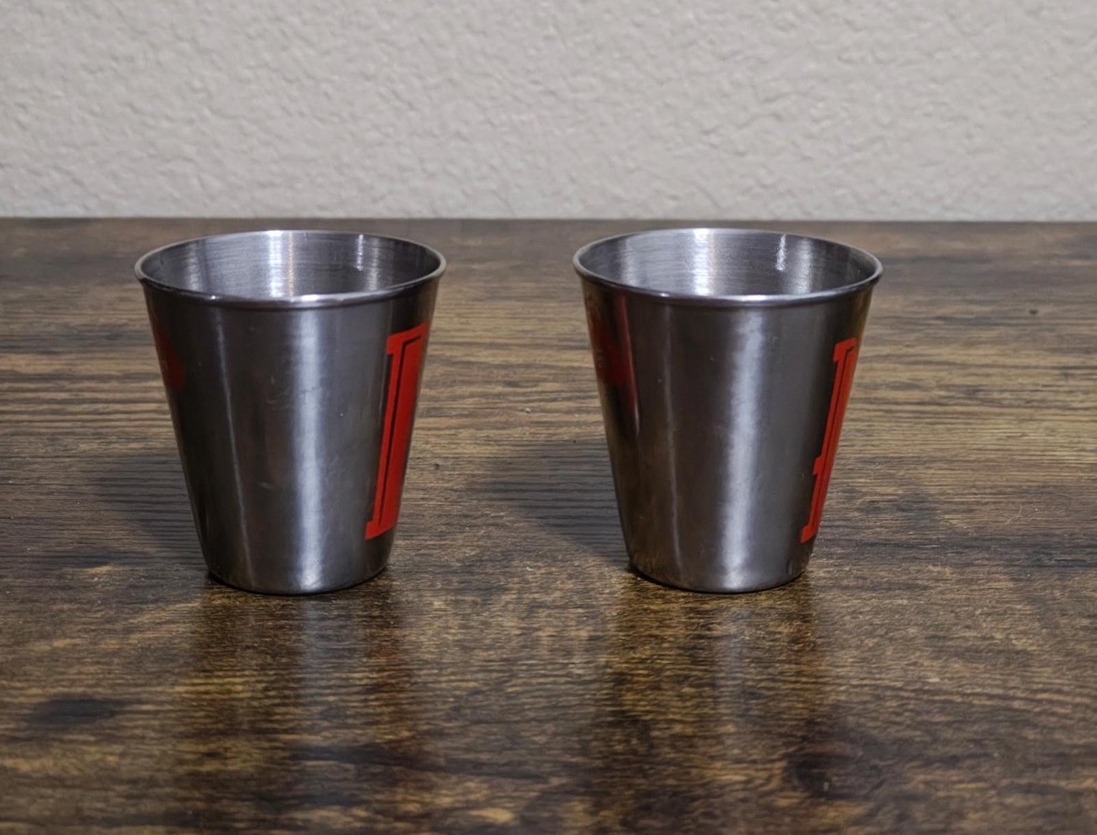 Pair of stainless steel shot glasses shown from the side with red vertical letters