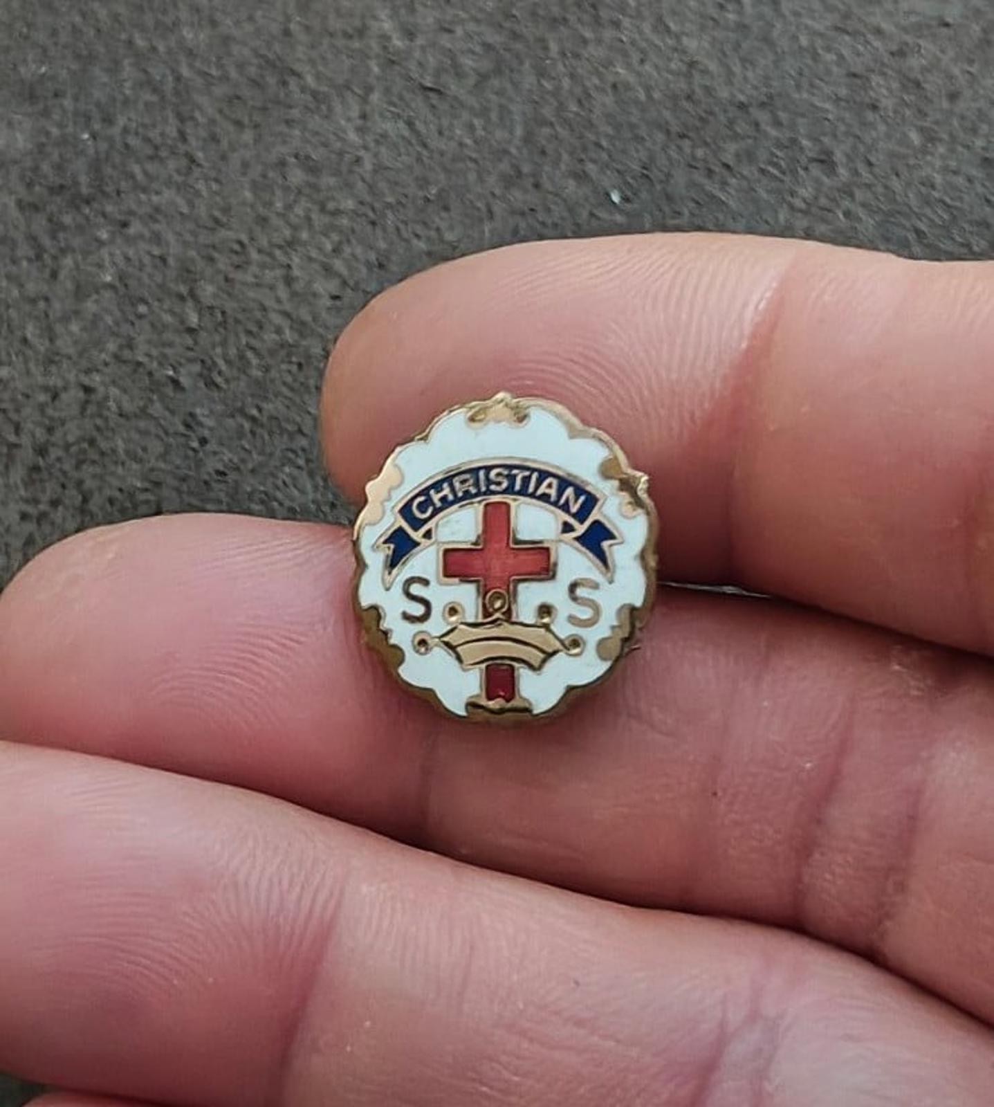 Small round Christian enamel pin in hand, gold rim with red cross and “S.O.S.”