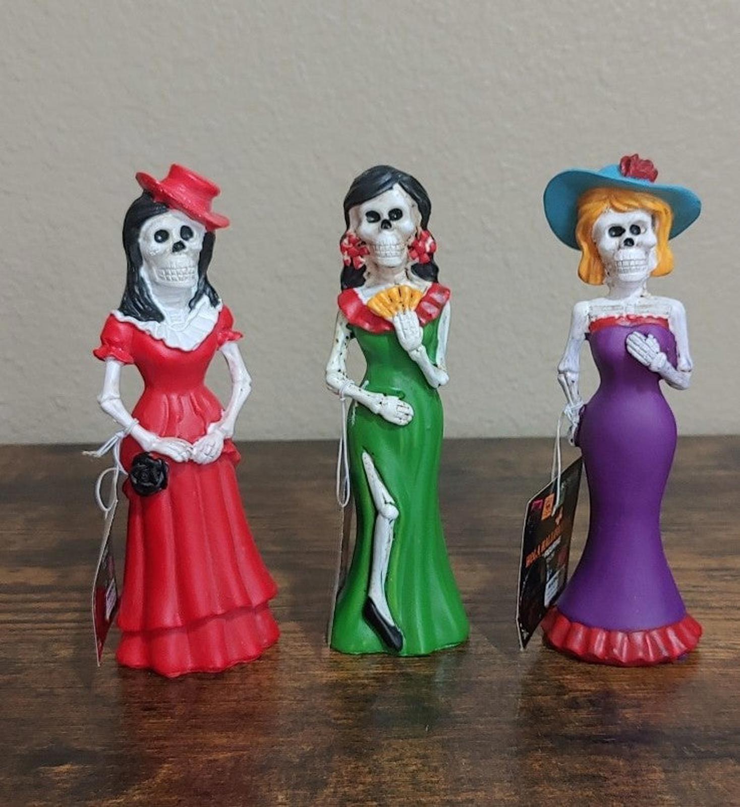 Three Catrina figurines standing side by side, red, green, and purple dresses