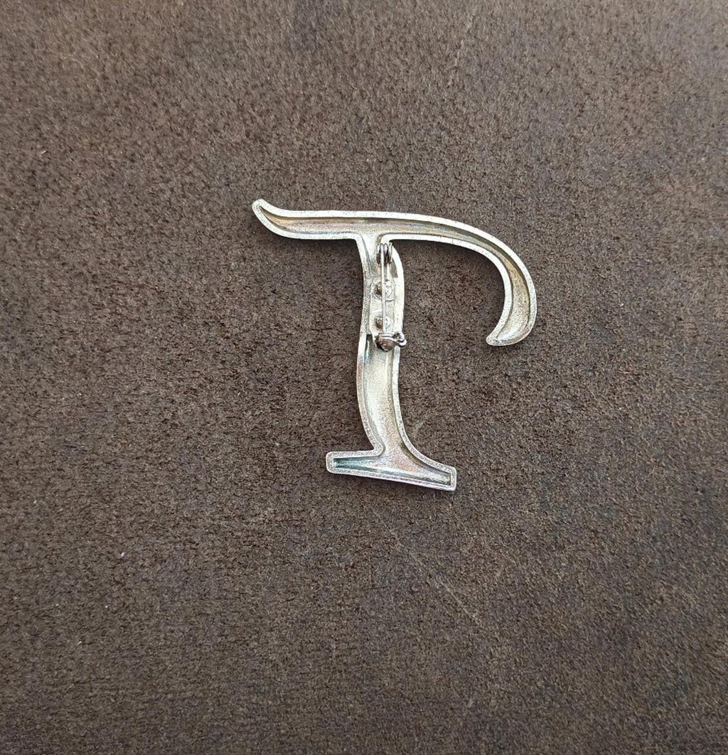 Silver-tone letter T pin/brooch with visible pin backing and clasp detail.