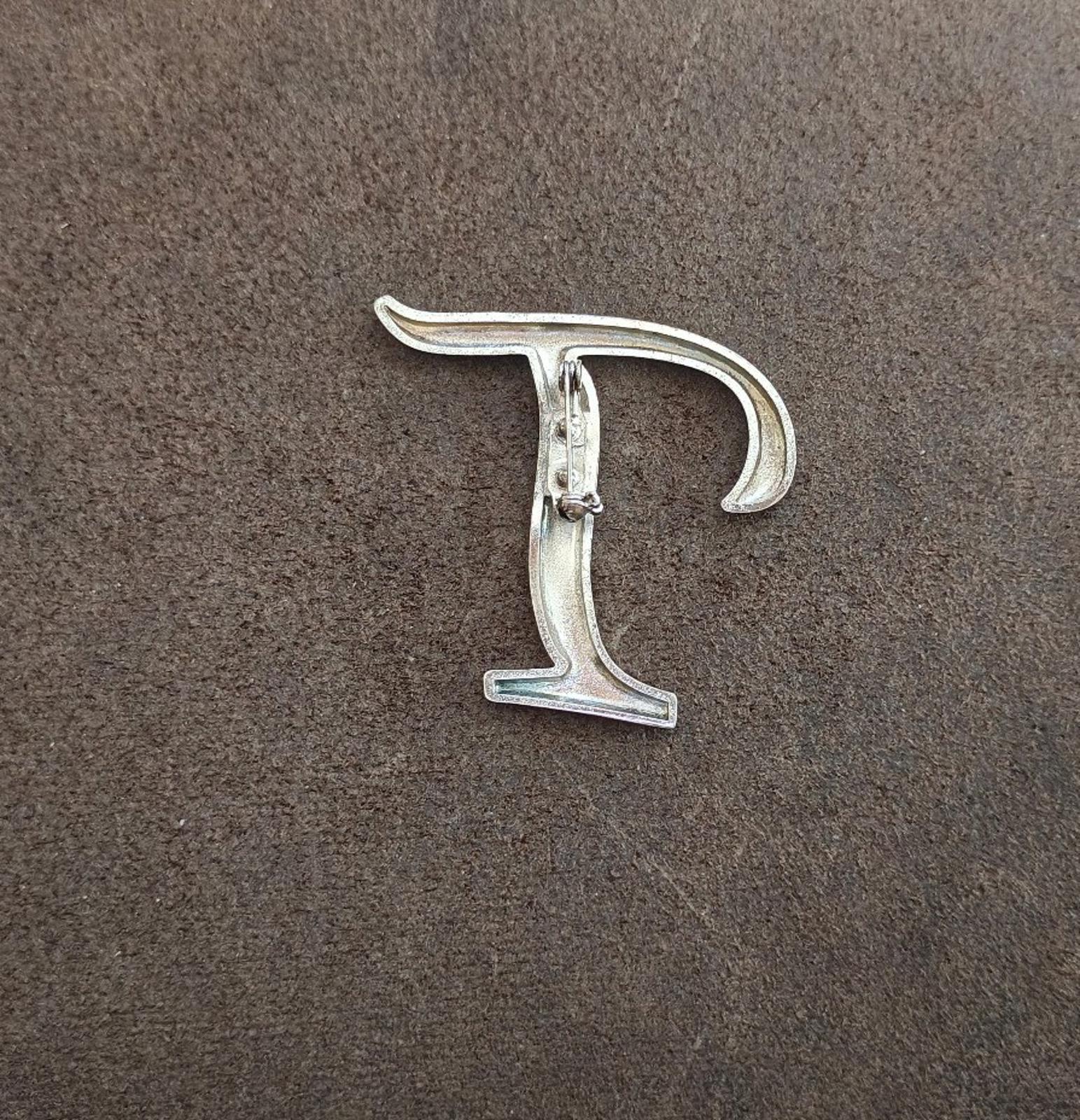 Silver-tone letter T pin/brooch with visible pin backing and clasp detail.