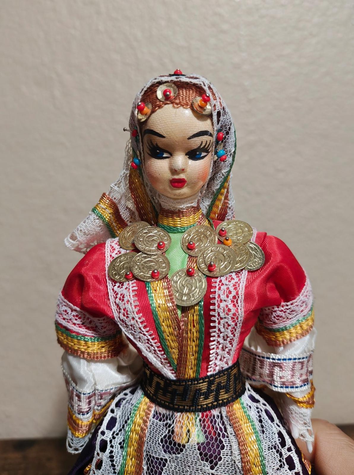 Female folk doll face with jeweled headscarf, red gown, and gold coin chest ornaments