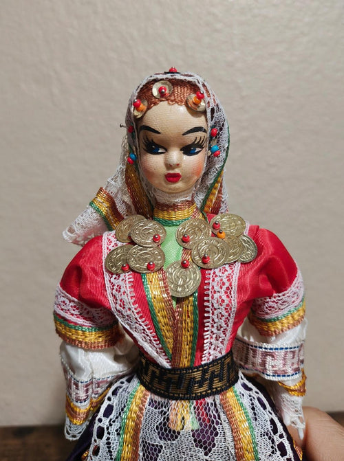 Female folk doll face with jeweled headscarf, red gown, and gold coin chest ornaments