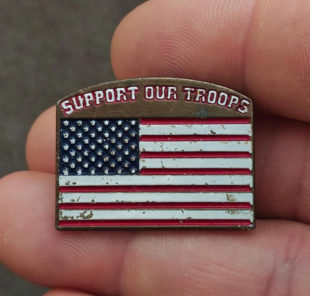 Gold-tone enamel pin in hand with “Support Our Troops” and U.S. stars-and-stripes flag