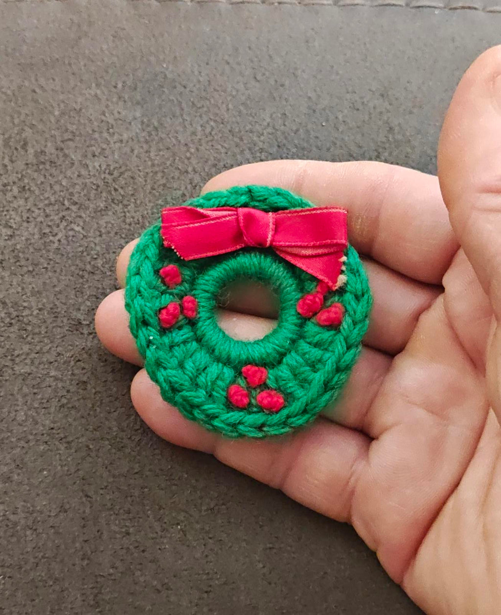 Green knitted wreath brooch with pink bow and red berry clusters, hand-held front view