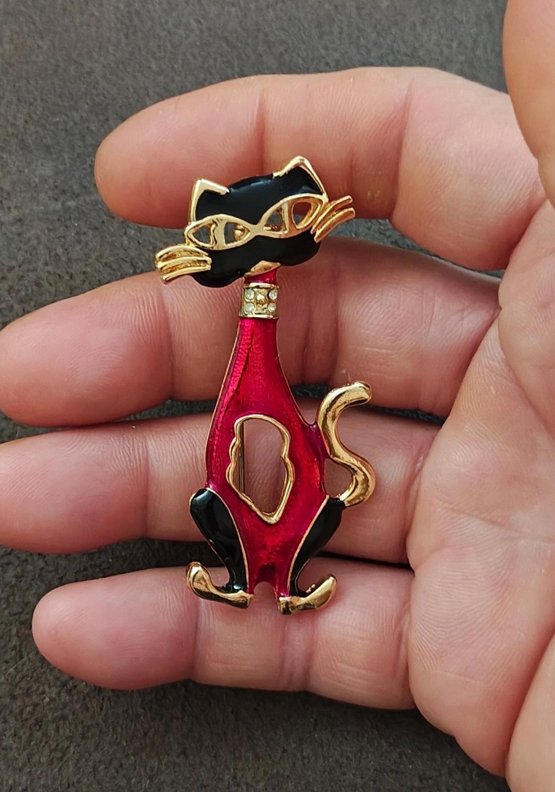 Red and black enamel cat mask brooch with gold-tone whiskers, held between fingers.