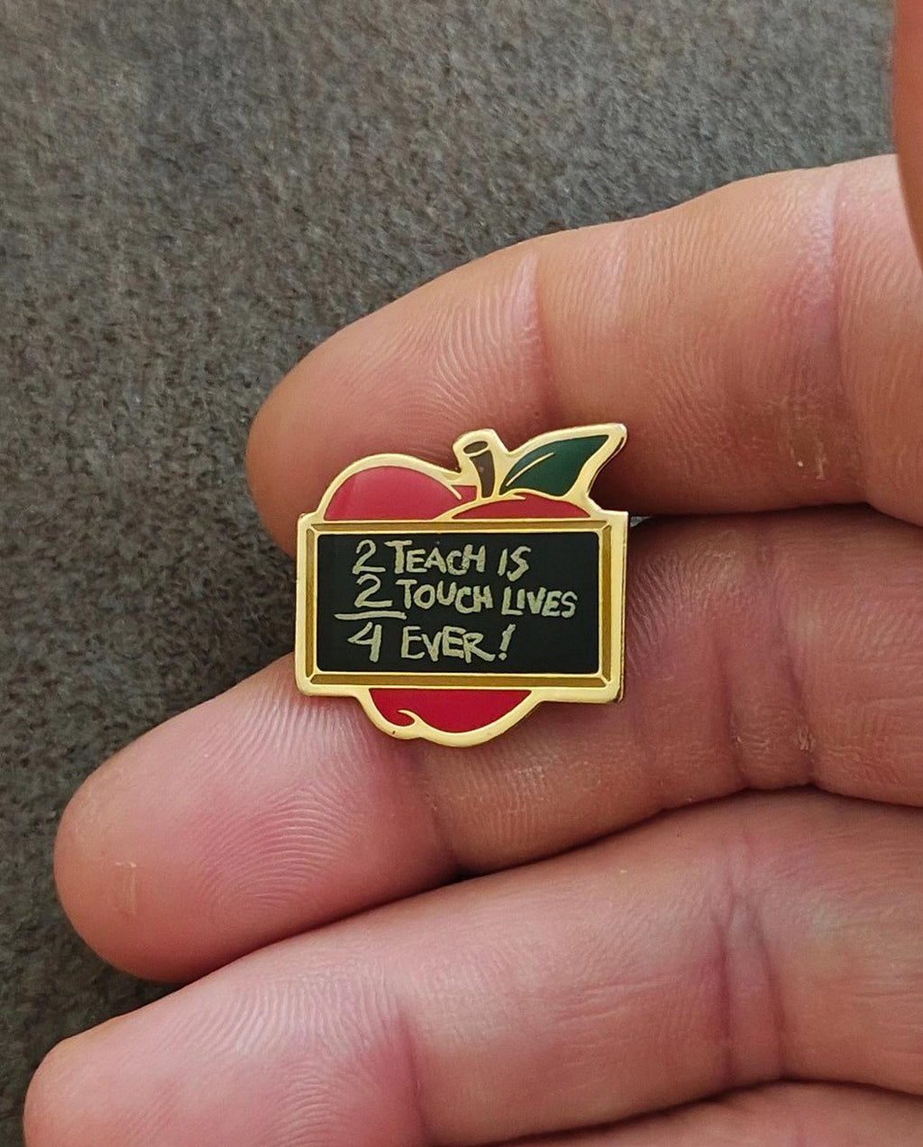 Gold-tone apple-shaped teacher pin with red enamel, green leaf, and black quote panel in hand
