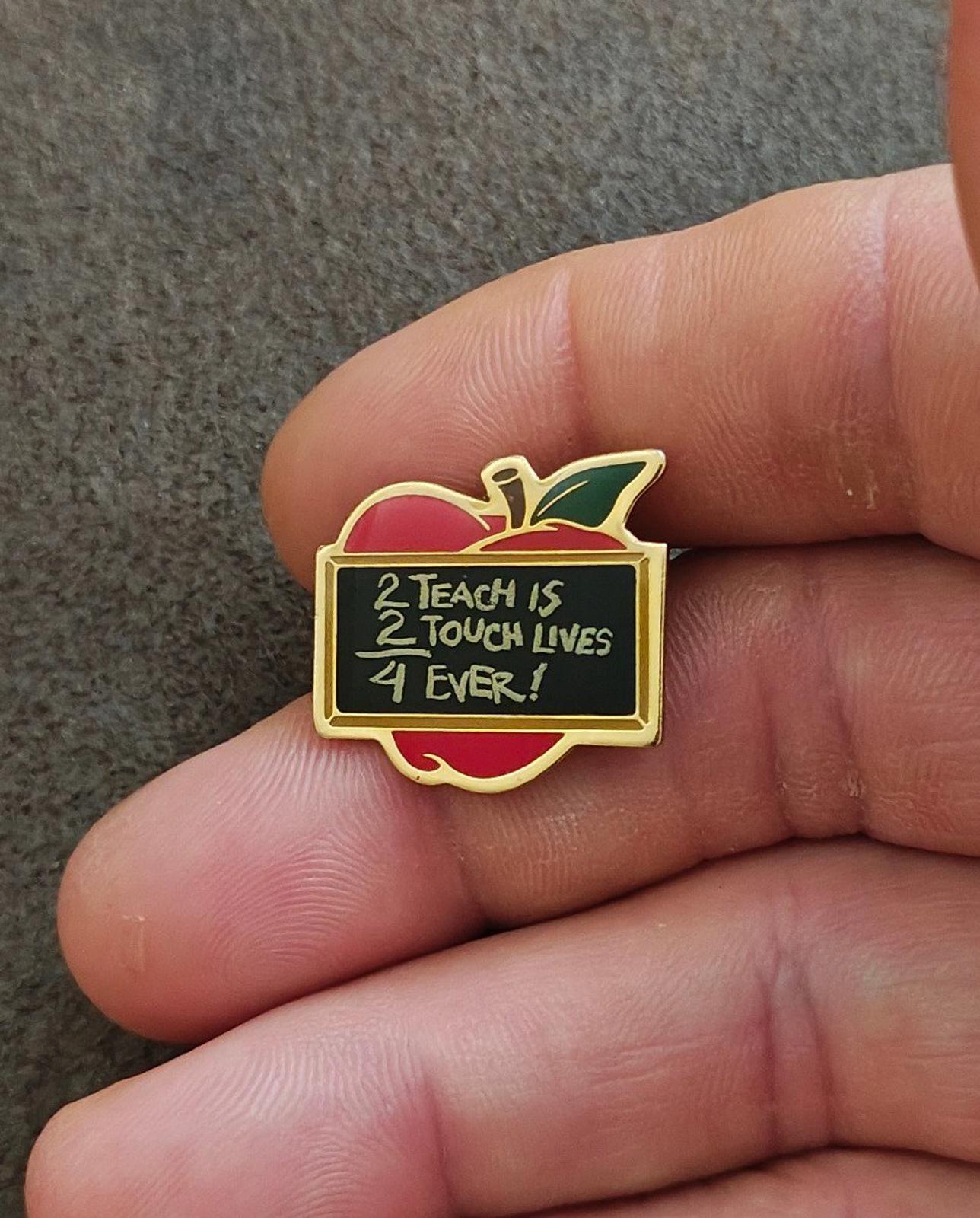 Gold-tone apple-shaped teacher pin with red enamel, green leaf, and black quote panel in hand